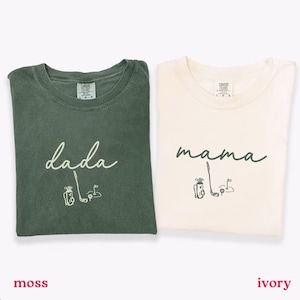 May include: Two t-shirts, one moss green and one ivory, are displayed. The moss green shirt says "dada" with a golf-themed graphic. The ivory shirt says "mama" with a golf-themed graphic. Both shirts are short-sleeved.