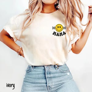 May include: Ivory-colored t-shirt with a smiley face graphic and the word "MAMA" in black. The smiley face is yellow with a black and white checkered pattern on each side. The word "ivory" is printed in black at the bottom left.