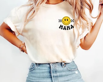 May include: Ivory-colored t-shirt with a smiley face graphic and the word "MAMA" in black. The smiley face is yellow with a black and white checkered pattern on each side. The word "ivory" is printed in black at the bottom left.