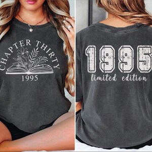 May include: Dark gray t-shirt with the text "CHAPTER THIRTY 1995" above an open book with floral illustrations on the front. The back of the shirt displays "1995 limited edition" in a collegiate style.