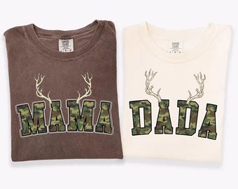 May include: Two t-shirts, one in espresso and the other in ivory, are shown. Each shirt has the words "MAMA" and "DADA" in a camouflage pattern, with a deer antler design above. The colour names are displayed at the bottom of the image.