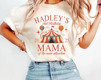 Pode incluir: Camiseta marfim com um design de tema circense. O design inclui uma tenda de circo listrada em vermelho e branco, balões de ar quente e o texto "HADLEY'S 1st Birthday" e "MAMA of the main attraction". Os números "20" e "25" também estão incluídos.