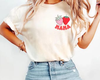 May include: Ivory-colored t-shirt featuring a strawberry design with the word "MAMA" in red. The shirt has rolled-up sleeves and is paired with blue jeans. The word "ivory" is printed at the bottom left.
