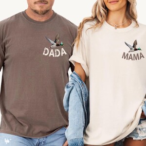 Puede incluir: Dos camisetas de manga corta, una marr&oacute;n y otra crema, cada una con un gr&aacute;fico de un pato volando. La camiseta marr&oacute;n tiene la palabra "DADA" impresa debajo del pato, mientras que la camiseta crema dice "MAMA".