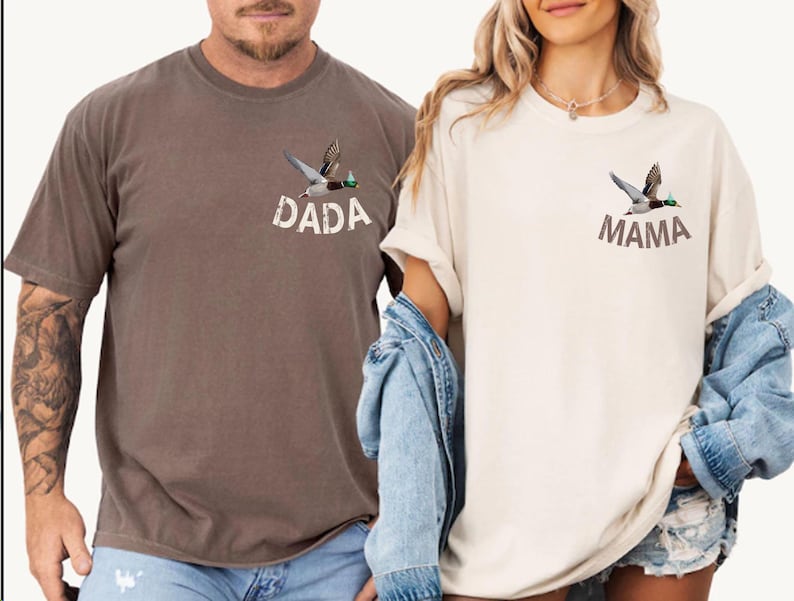 Puede incluir: Dos camisetas de manga corta, una marr&oacute;n y otra crema, cada una con un gr&aacute;fico de un pato volando. La camiseta marr&oacute;n tiene la palabra "DADA" impresa debajo del pato, mientras que la camiseta crema dice "MAMA".
