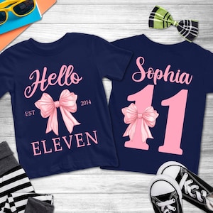 May include: Two navy blue t-shirts with pink text and bow graphics. One shirt says "Hello Eleven" with a bow and "EST 2014". The other says "Sophia 11" with a bow and the number 11.