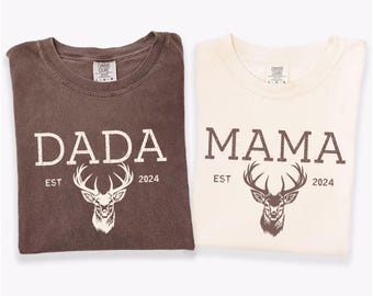 May include: Two folded t-shirts, one espresso and one ivory. The espresso shirt reads "DADA" with a deer graphic and "EST 2024". The ivory shirt reads "MAMA" with a deer graphic and "EST 2024".