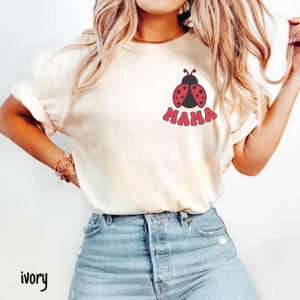 May include: Ivory-colored t-shirt featuring a red and black ladybug design above the word "MAMA" in red, retro-style lettering. The shirt is paired with blue denim jeans. The word "ivory" is printed at the bottom left.