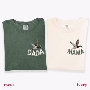 Puede incluir: Dos camisetas dobladas, una verde musgo y otra marfil. Cada camiseta presenta un gr&aacute;fico de un pato en vuelo sobre las palabras "DADA" y "MAMA". Las camisetas est&aacute;n etiquetadas como "moss" e "ivory" en la parte inferior.