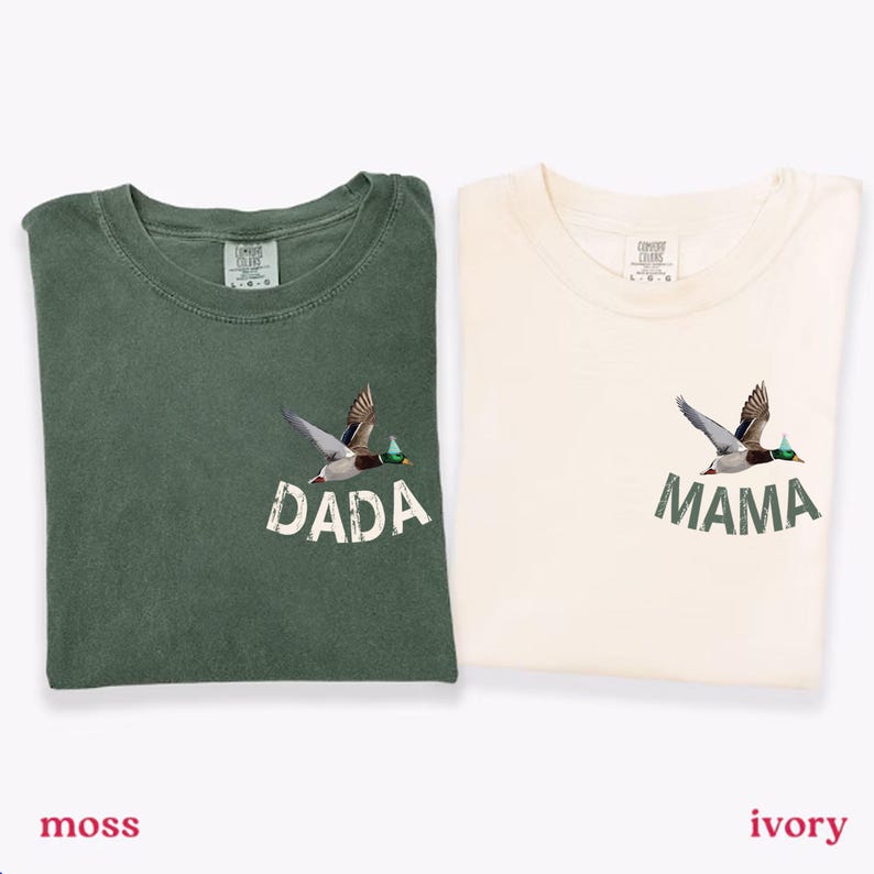 Puede incluir: Dos camisetas dobladas, una verde musgo y otra marfil. Cada camiseta presenta un gr&aacute;fico de un pato en vuelo sobre las palabras "DADA" y "MAMA". Las camisetas est&aacute;n etiquetadas como "moss" e "ivory" en la parte inferior.