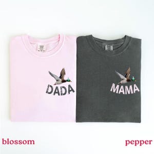 Puede incluir: Dos camisetas, una rosa flor y otra gris pimienta, cada una con un dise&ntilde;o de pato volador. La camiseta rosa tiene la palabra "DADA" y la camiseta gris tiene la palabra "MAMA" impresa debajo.