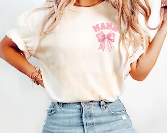 May include: Ivory-colored t-shirt with rolled-up sleeves. The shirt features the word "MAMA" in pink bubble letters above a pink bow graphic. The word "ivory" is printed in black at the bottom left.