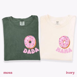 May include: Two t-shirts, one moss green and one ivory, each featuring a pink donut graphic with sprinkles. The moss green shirt has the word "DADA" below the donut, while the ivory shirt says "MAMA".