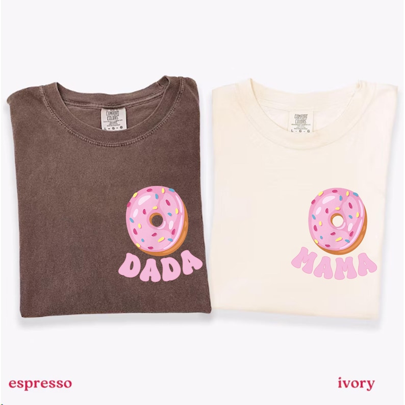 May include: Two t-shirts, one espresso and one ivory, each with a pink donut graphic. The espresso shirt has the word "DADA" in pink, while the ivory shirt says "MAMA". Both shirts are folded.