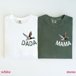 Puede incluir: Dos camisetas, una blanca y otra verde musgo, cada una con un gr&aacute;fico de pato. La camiseta blanca tiene la palabra "DADA" y la camiseta verde tiene la palabra "MAMA" impresa debajo de la ilustraci&oacute;n del pato. Las camisetas son de material suave.