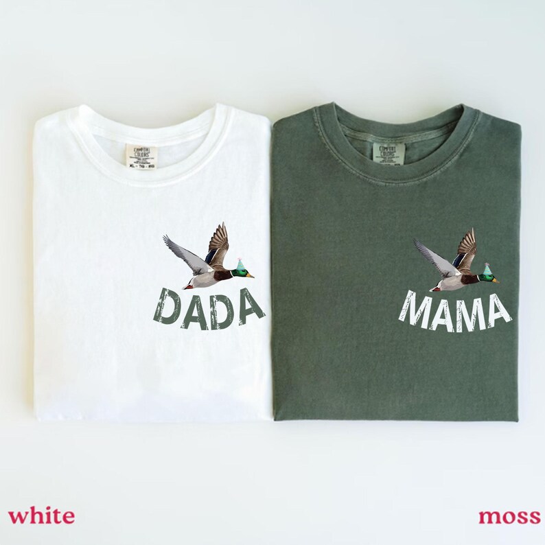 Puede incluir: Dos camisetas, una blanca y otra verde musgo, cada una con un gr&aacute;fico de pato. La camiseta blanca tiene la palabra "DADA" y la camiseta verde tiene la palabra "MAMA" impresa debajo de la ilustraci&oacute;n del pato. Las camisetas son de material suave.