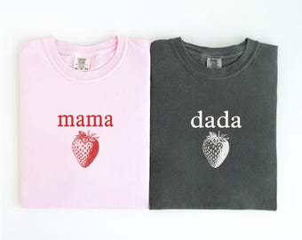 May include: Two folded t-shirts, one light pink and one dark gray. The pink shirt has the word "mama" and a red strawberry graphic. The gray shirt has the word "dada" and a white strawberry graphic. The word "blossom" is below the pink shirt and "pepper" is below the gray shirt.