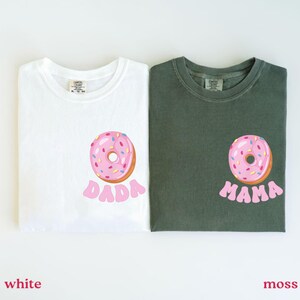 May include: Two t-shirts, one white and one moss green, each featuring a pink donut graphic with sprinkles. The white shirt has the word "DADA" below the donut, and the green shirt says "MAMA".