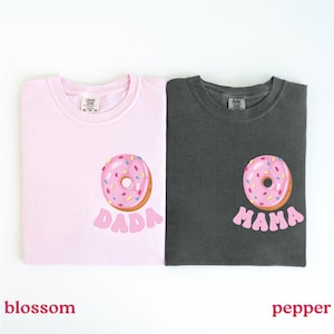 May include: Two t-shirts, one blossom pink and one pepper gray, each featuring a donut graphic. The pink shirt has the word "DADA" below the donut, while the gray shirt says "MAMA". The donuts have pink frosting and colorful sprinkles.