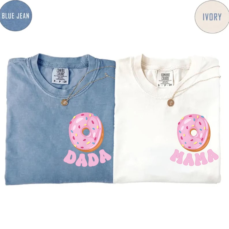 May include: Two t-shirts, one blue and one ivory, each with a pink donut graphic and text. The blue shirt says "DADA" and the ivory shirt says "MAMA". Both shirts have a gold necklace.