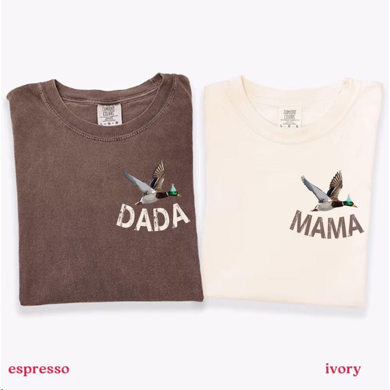 Puede incluir: Dos camisetas, una espresso y otra marfil, cada una con un gr&aacute;fico de pato volador. La camiseta espresso tiene la palabra "DADA" impresa debajo del pato, mientras que la camiseta marfil dice "MAMA". Ambas camisetas tienen cuello redondo.