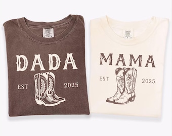 May include: Two t-shirts, one espresso and one ivory, are displayed. The espresso shirt reads "DADA" above a cowboy boot graphic, with "EST 2025" below. The ivory shirt says "MAMA" with a similar boot graphic and "EST 2025".