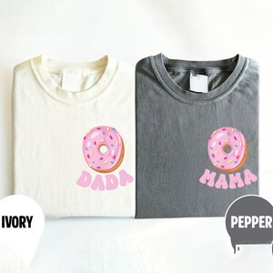 May include: Two folded t-shirts, one ivory and one pepper gray, each with a pink donut graphic. The ivory shirt has the word "DADA" in pink, while the gray shirt says "MAMA".