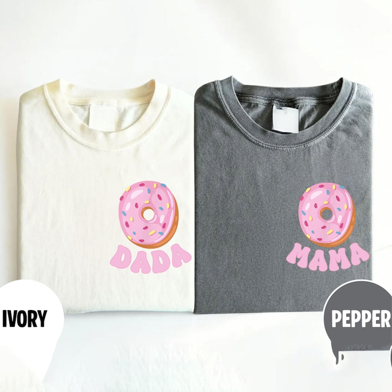 May include: Two folded t-shirts, one ivory and one pepper gray, each with a pink donut graphic. The ivory shirt has the word "DADA" in pink, while the gray shirt says "MAMA".