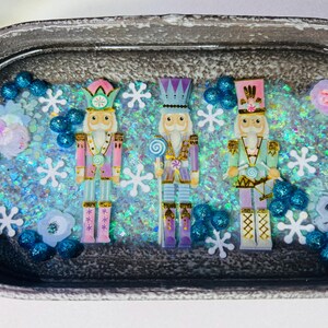 May include: Three colorful nutcracker figurines in a decorative tray with glitter, snowflakes, and floral accents. The nutcrackers are dressed in pink, purple, and green uniforms with gold details. The tray has a silver-gray finish.