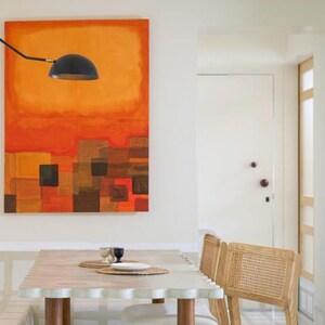 May include: An abstract painting with orange and brown hues hangs above a table with a scalloped edge. A black lamp is positioned above the table. Two chairs with woven backs are visible.