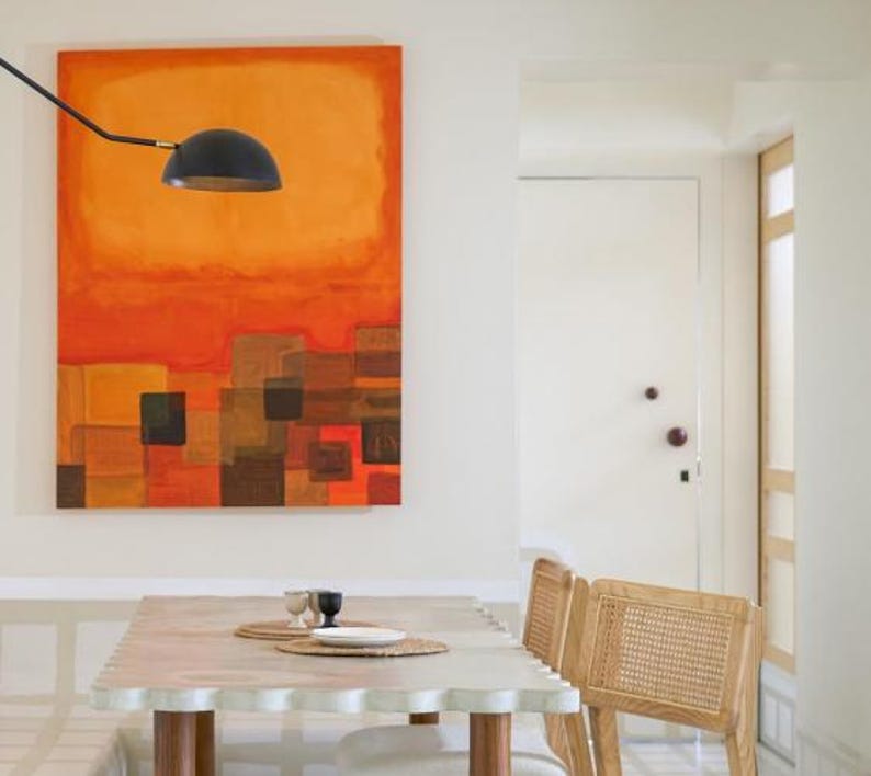 May include: An abstract painting with orange and brown hues hangs above a table with a scalloped edge. A black lamp is positioned above the table. Two chairs with woven backs are visible.