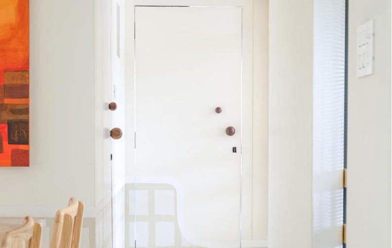 May include: A bright white door with two wooden knobs and a dark lock. The door is in a white hallway with a painting on the left and a window on the right. The door is closed.