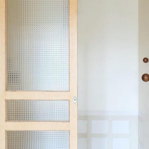 May include: A light-colored wooden door with three glass panels. The glass has a textured, grid-like pattern. The door is partially open, revealing a white wall and a glimpse of a window.