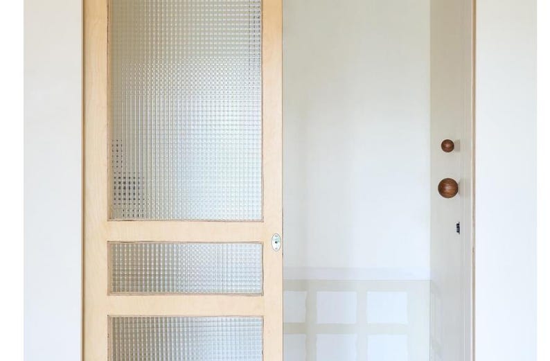 May include: A light-colored wooden door with three glass panels. The glass has a textured, grid-like pattern. The door is partially open, revealing a white wall and a glimpse of a window.