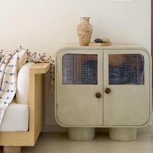 May include: A cream-colored cabinet with two doors and textured glass panels. The cabinet has rounded edges, wooden knobs, and sits on cylindrical legs. A decorative vase and stones rest on top. A cream-colored sofa with a blue patterned throw is to the left.