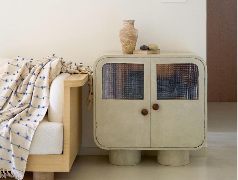 May include: A cream-colored cabinet with two doors and textured glass panels. The cabinet has rounded edges, wooden knobs, and sits on cylindrical legs. A decorative vase and stones rest on top. A cream-colored sofa with a blue patterned throw is to the left.