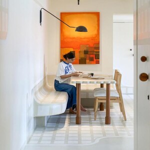 May include: A dining area with a white and beige bench, a light-colored wooden table, and a wooden chair. An abstract orange and brown painting hangs on the wall. A person is sitting at the table.