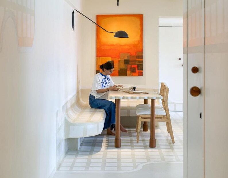 May include: A dining area with a white and beige bench, a light-colored wooden table, and a wooden chair. An abstract orange and brown painting hangs on the wall. A person is sitting at the table.