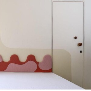 May include: A bedroom interior featuring a bed with a white comforter and a headboard with a wavy design in shades of red and pink. The wall has a matching design in beige and white, and a white door with wooden knobs.