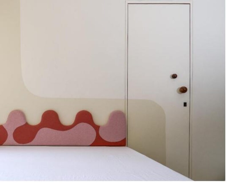 May include: A bedroom interior featuring a bed with a white comforter and a headboard with a wavy design in shades of red and pink. The wall has a matching design in beige and white, and a white door with wooden knobs.