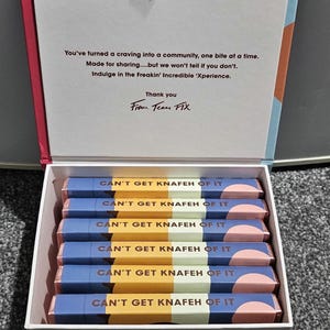Könnte beinhalten: Eine weiße rechteckige Schachtel mit sechs Schokoriegeln in blau-gelb-rosa Verpackungen, jeweils beschriftet mit CAN'T GET KNAFEH OF IT. Auf der Innenseite des Deckels steht: You've turned a craving into a community, one bite at a time. Thank you From Team FIX.