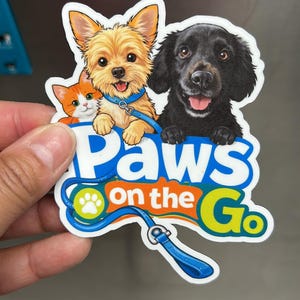 Puede incluir: Una colorida pegatina con un Yorkshire Terrier dorado, un Labrador negro y un gato naranja y blanco. La pegatina tiene el texto "Paws on the Go" con una huella de pata y un diseño de correa. La pegatina está sujeta por una mano.