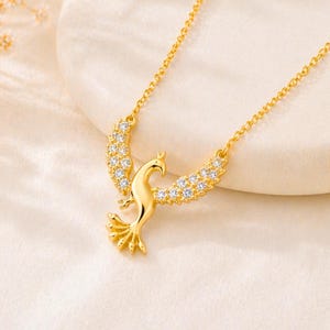 May include: A gold-colored necklace featuring a phoenix pendant. The phoenix has outstretched wings adorned with small, clear gemstones. The necklace chain is delicate, and the pendant is centered. The background is a soft, neutral color.