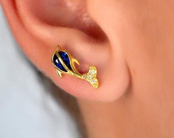 Puede incluir: Un pendiente de botón dorado con forma de delfín, que presenta un cuerpo de esmalte azul intenso y una cola acentuada con pequeñas piedras claras y brillantes. El pendiente está colocado en el lóbulo, mostrando su diseño detallado y pulido.