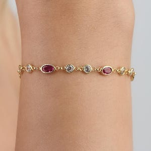 Puede incluir: Una delicada pulsera de cadena dorada que presenta una alternancia de piedras preciosas rojas de forma ovalada y piedras blancas transparentes redondas engastadas en biseles metálicos individuales, unidas por pequeños eslabones dorados.