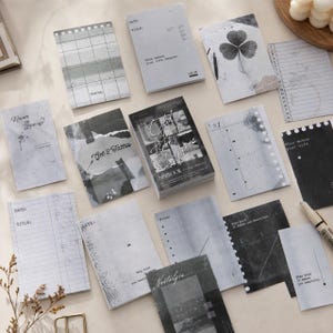 Op de afbeelding: Een verzameling rechthoekige papieren memoblaadjes en een kleine stapel kaarten in grijstinten, zwart en wit. De items bevatten diverse gedrukte ontwerpen, waaronder rasterpatronen, bloemillustraties en teksten zoals "Never Give Up", "Get 2 Tattoos", "Nostalgia" en "Stay Kind It Makes You Beautiful".