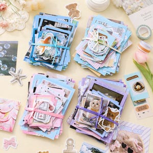 May include: Four stacks of assorted paper ephemera and stickers tied with ribbons. The stacks feature blue, pink, and purple color themes with various printed images including a goldfish, a cat, and text like I GIVE and COOL CAT.