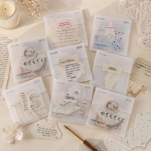 May include: A collection of eight translucent envelopes labeled MO-CARD, each containing assorted paper ephemera, vintage-style poetry snippets, and decorative text labels. A gold-toned fountain pen and a small paper cutout lie nearby.