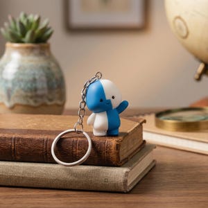 Peut inclure: Un porte-clés figurine bleu et blanc avec une chaîne et un anneau argentés est posé sur une pile de vieux livres. La figurine est dans une pose de salut. Un vase décoratif avec une plante succulente et un globe sont en arrière-plan.