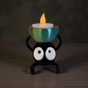Puede incluir: Una figura negra caprichosa sostiene un cuenco turquesa y dorado con una vela LED parpadeante. La figura tiene ojos blancos grandes y un cuerpo redondo. La vela emite un cálido resplandor anaranjado, creando un ambiente acogedor.
